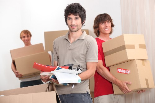 A group of three young men participating in a house move inside a bright, well-lit room with neutral-colored walls and curtains. The man in the foreground has dark, curly hair, light skin, and is wearing a light grey shirt. He is holding a stack of documents or leaflets, along with a pair of safety gloves, and is looking directly at the camera with a slight smile. Behind him, to his left, another young man with blonde hair and light skin is holding a large cardboard box. To his right, a third young man with curly brown hair and light skin is smiling while carrying two stacked cardboard boxes, one marked with a red “Fragile” sticker. The boxes vary in size and are made of brown corrugated cardboard, some sealed with packing tape. The setting indicates an indoor home environment, likely a living room, with minimal visible furniture and a clean, uncluttered space, suggesting an organized house removal process. The individuals’ casual clothing and the presence of packing materials reflect a typical household move facilitated by a professional removals service, aligned with the context of Chelsea Man with Van’s house removal offerings in Chelsea and nearby areas.