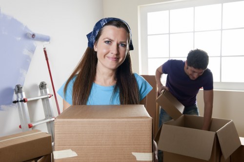 A woman with long brown hair, wearing a blue top and a patterned headscarf, stands in a well-lit room holding a large cardboard box in front of her. Behind her, a man with dark curly hair, dressed in a navy blue shirt and dark trousers, is bent over, placing or inspecting a cardboard box near a cream-colored wall with a large window that allows natural light to illuminate the space. The room appears to be part of a residential property, possibly in Chelsea, with a staircase on the left and several packing boxes of various sizes scattered around, some closed and others open. The boxes are made of brown cardboard, with clear packing tape securing some of them. The environment suggests an ongoing house move or packing process typical of professional removals, with a focus on careful handling and organization of household belongings, supported by the presence of multiple boxes and minimal furnishings visible in the neutral-toned interior.