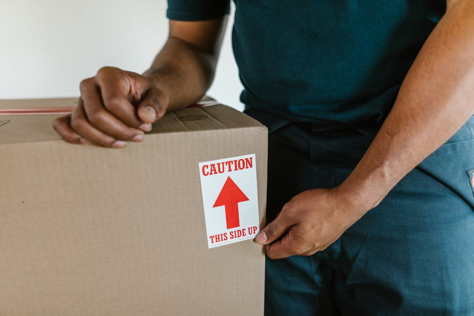 A person with dark skin holding an open cardboard box labelled 'FRAGILE' with red and white sticker, in a packing area surrounded by other sealed cardboard boxes. The individual is wearing a maroon to