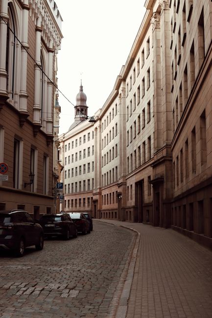 A narrow street lined with multi-storey buildings featuring colorful facades, with a sign for a pub hanging from one building and flower baskets hanging beside it. On the street, there are two deliver