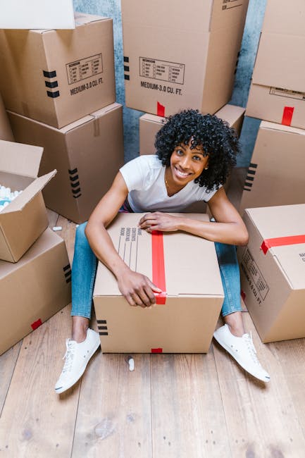 A young woman with curly dark hair, wearing a white T-shirt, blue jeans, and white sneakers, sitting on a wooden floor surrounded by numerous cardboard boxes of varying sizes, some sealed with red and